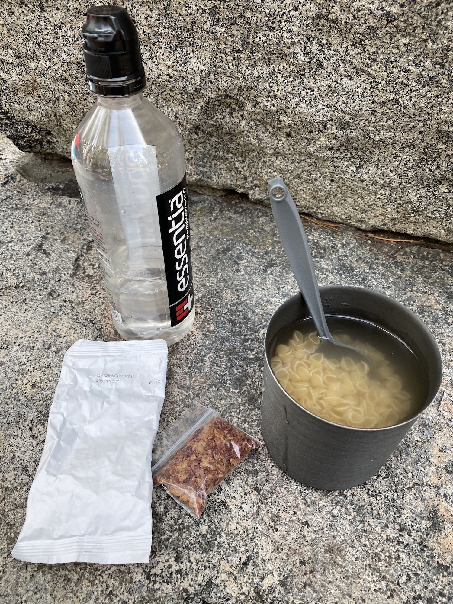 A backpacking pot with macaroni and water. The cheese and bacon packets are next to it