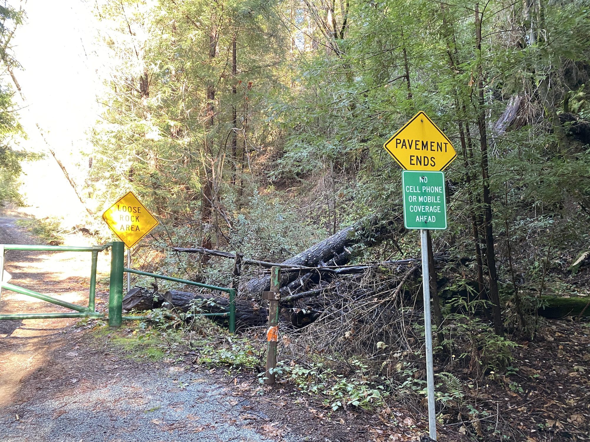 A locked gate with signs warning of loose rocks, lack of pavement, and no cell phone signal.