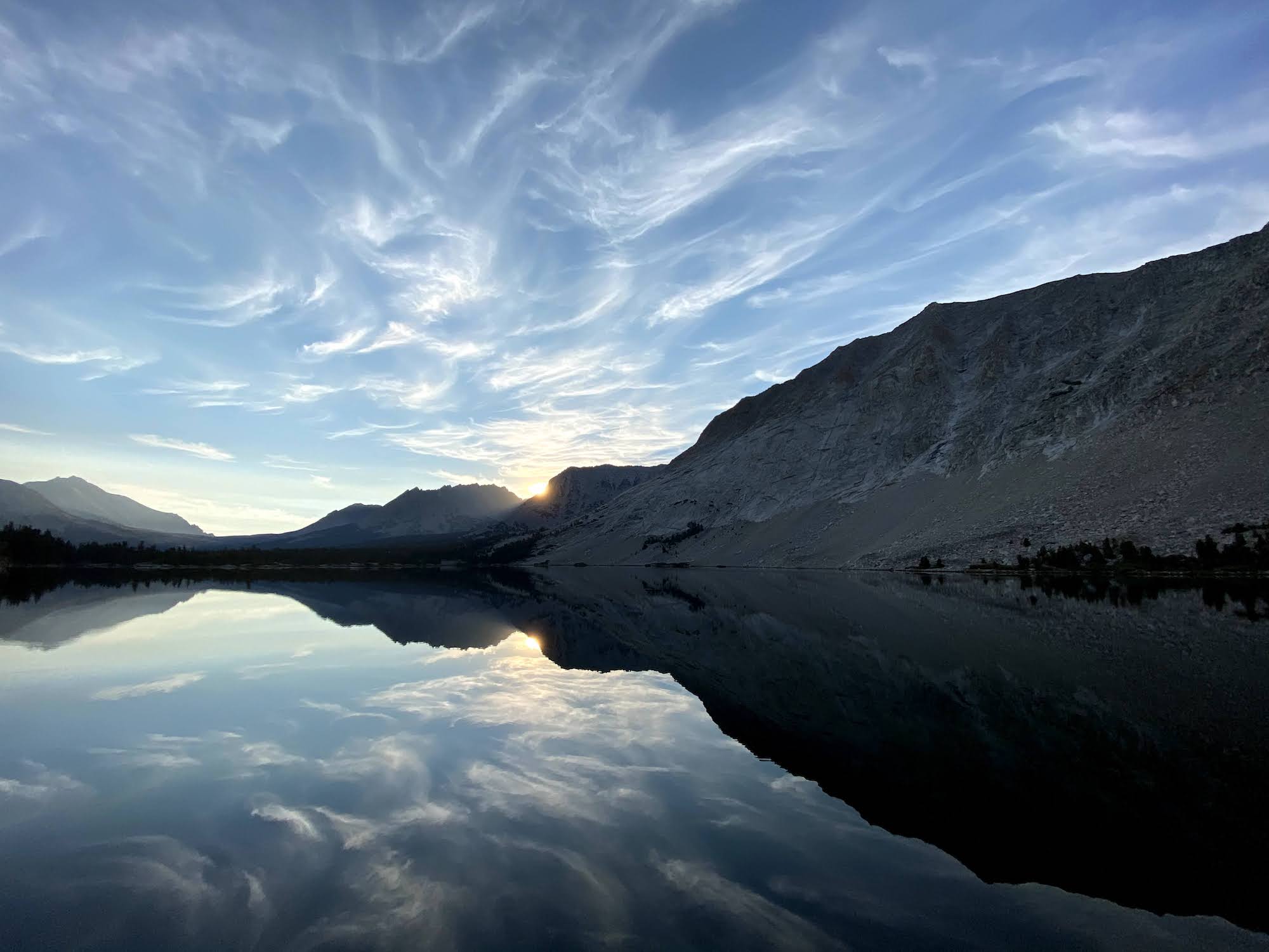 The morning sun peeking above the mountains behind a lake.