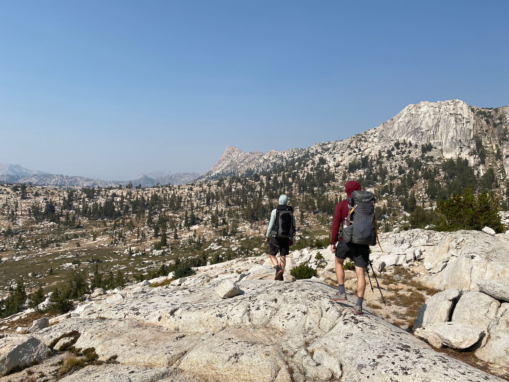 Two backpackers walking in a high alpine terrain with granite and small trees.