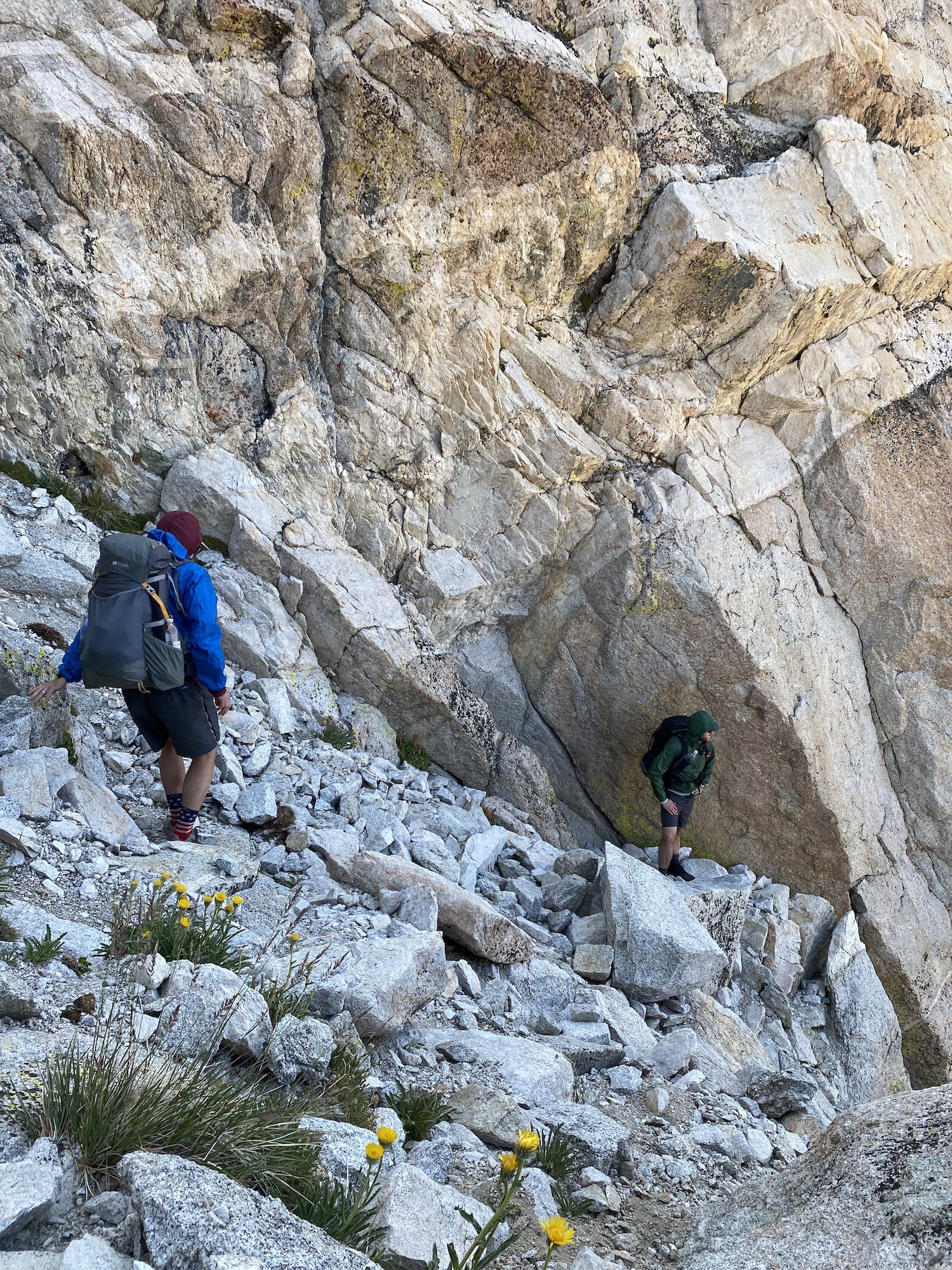 Two backpackers on steep talus with a sheer granite wall behind them.