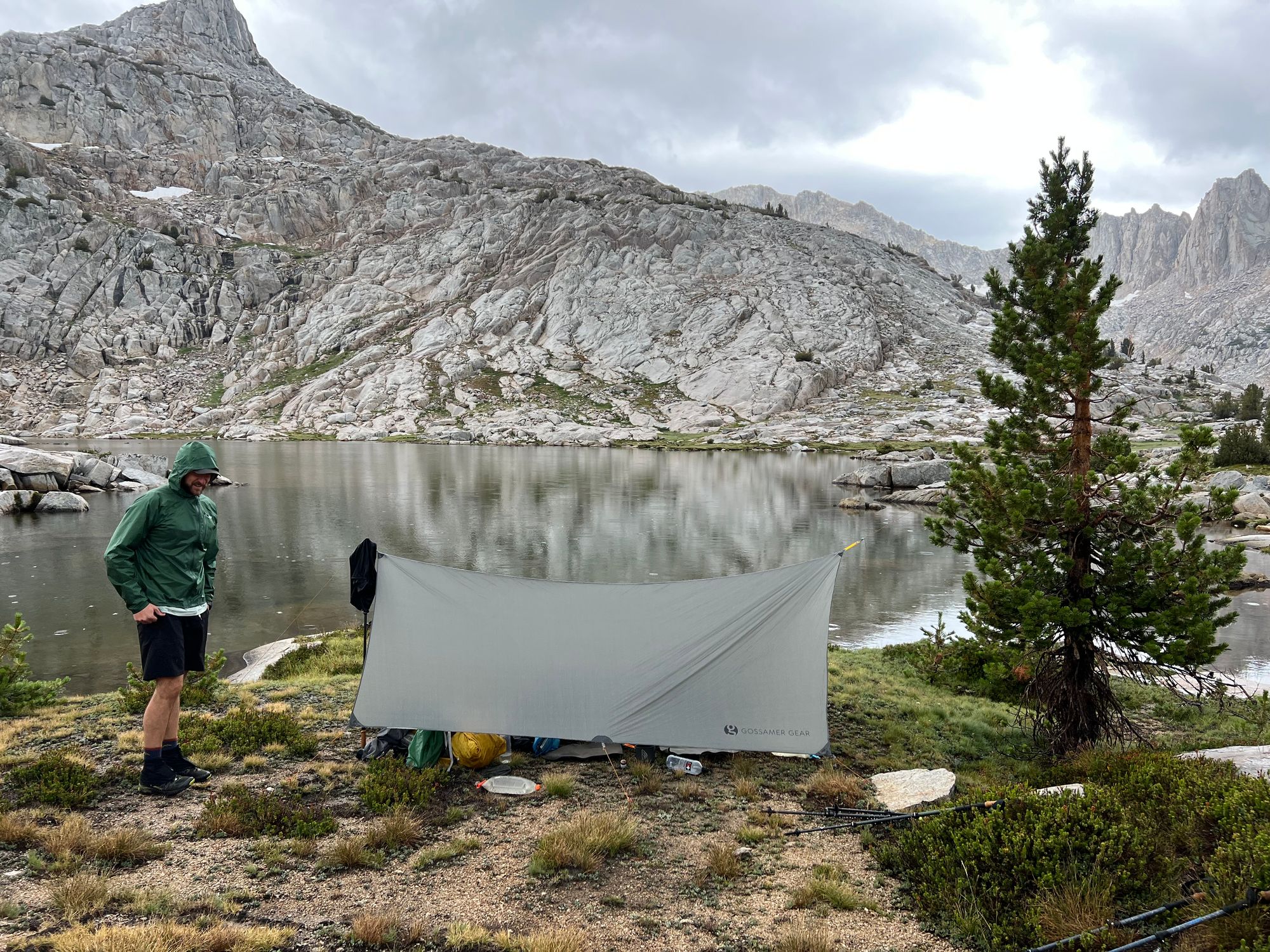 A man in a wet rain jacket standing next to a tarp set up in front of a lake.