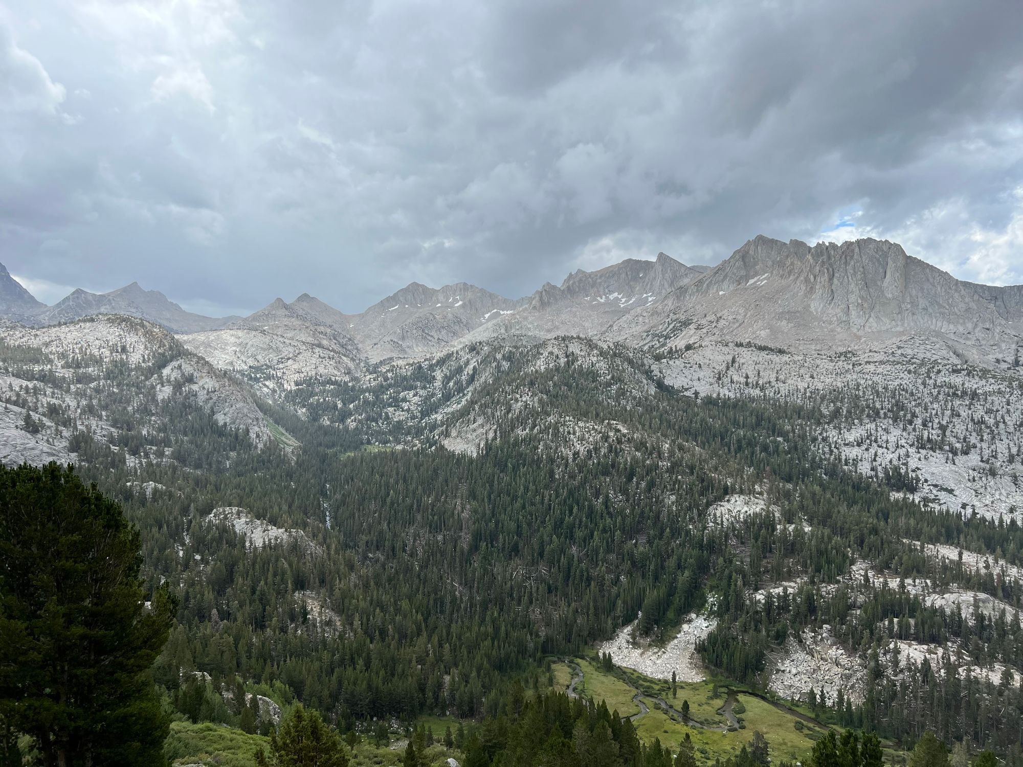Dark clouds over bare mountain peaks. A meadow with a stream in the foreground.